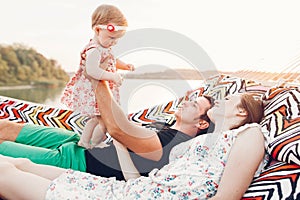 Strong young father holding his baby daughter in hands with happy hipster mother, smiling family relaxing in a hammock on a beach