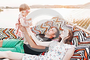 Strong young father holding his baby daughter in hands with happy hipster mother, smiling family relaxing in a hammock on a beach