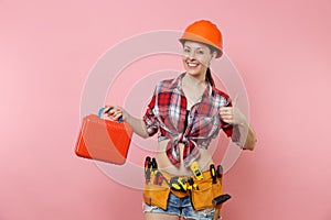 Strong young handyman woman in orange helmet, plaid shirt, denim shorts, kit tools belt full of instruments, toolbox