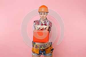 Strong young handyman woman in orange helmet, plaid shirt, denim shorts, kit tools belt full of instruments, toolbox