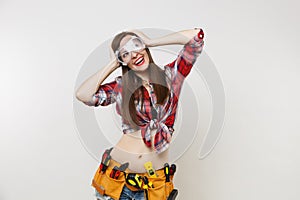 Strong young handyman woman in plaid top shirt, denim shorts, kit tools belt full of instruments in protective goggles