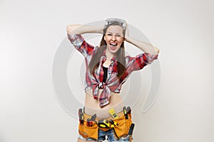 Strong young handyman woman in plaid top shirt, denim shorts, kit tools belt full of instruments in protective goggles