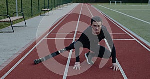 Strong young male sportsman with prosthetic running blades stretching limbs at sports field. Man with amputed leg doing