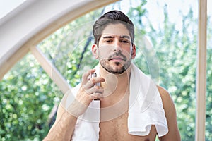 A stubble faced handsome man standing in the bathroom and puting cologne on his face