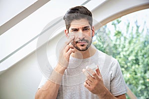 A stubbled face man is standing in front of the mirror in the bathroom and applying moisturizing cream to his face photo