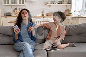 Stubborn son annoy mother at home: tired mom try to read message sit on sofa with naughty child