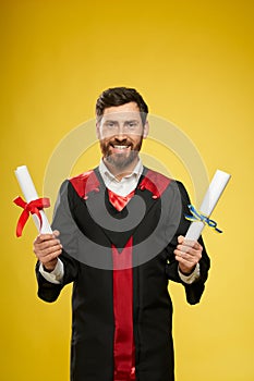 Student with beard standing, holding two diplomas.