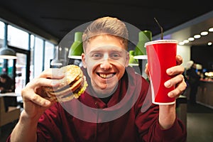 Student with a burger and a glass of cola against the background of a fast food restaurant