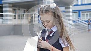 Student-centered elementary school in school uniform writes or draws something in a notebook near the school building.