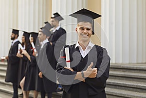 Student with a diploma in his hands stands on the steps of the university and looks into the camera.