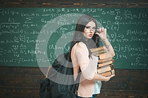 Student excellent accumulates knowledge. Girl with big backpack holds pile books, chalkboard background. Girl student