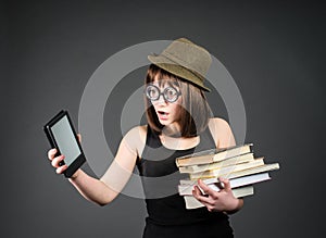 Student in funny glasses with old books in one hand and e-reader in another on grey background. Nerd girl is comparing