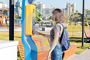 A student girl with a backpack presses the emergency terminal help button on a city street. The girl asks for help