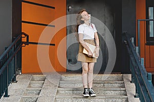 A student girl with a backpack is standing on the stairs near the house, clutching a notebook, enjoying the fresh air