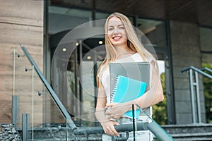 student girl holds folders, notebooks in hands and smiles on the background of the university building. Copy space