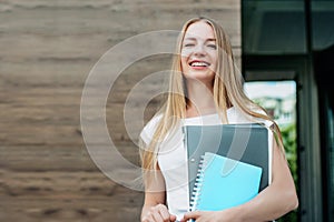 student girl holds folders, notebooks in hands and smiles on the background of the university building. Copy space
