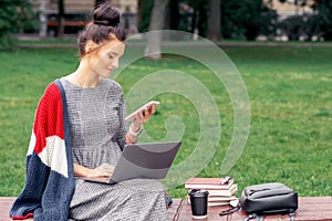Student girl is looking on smartphone with laptop on a wooden bench at park