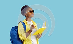 Student girl standing on blue background, holding pencil and notebook, writing essay and thinking