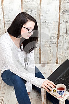 Student girl is wearing pair of glasses is studying on laptop while drinking from her mug.