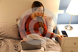 Student girl working on laptop on bed distracted by phone at the night time photo