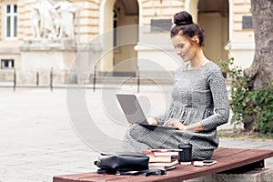 Student girl is working on laptop on bench near university building.