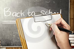 Student hands holding clipboard and writing in the classroom with piles of books and blackboard background