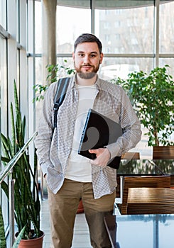 Student with laptop and backpack near window in reopen university campus. Caucasian teenager, confident bearded man carrying