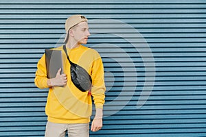Student with a laptop in his hands and a fanny pack around the neck standing against the background of a blue wall and looking