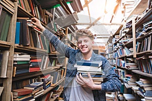 Student with a lot of books in his hands is in the library, takes books from the shelf and looks at the camera