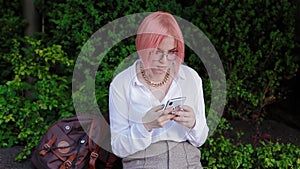 A student with pink hair puts a backpack on the parapet and takes out a mobile phone from her pocket