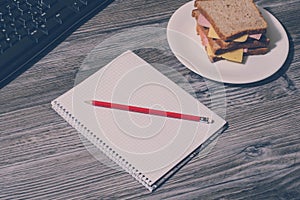 Student`s hard working. Tasty big sandwich on round plate, notebook with pencil, keyboard on gray wooden background. Vintage effe