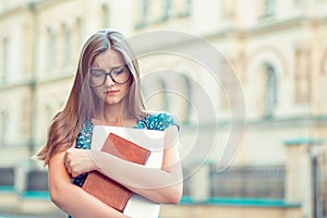 Student sad woman with glasses books, laptop in hand looking down in front of college