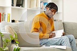 Student studying at home with laptop and books sitting on couch. Man preparing for college exams making notes.