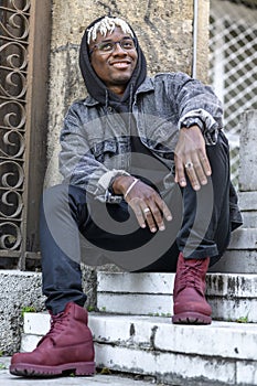 Student wear stylish fashion wear. Smiling African American man with dreadlocks sits on the steps near campus