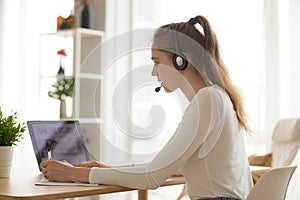 Student woman in headset writing notes, using laptop at workplace