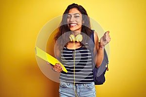 Student woman wearing bakcpack notebook headphones over isolated yellow background screaming proud and celebrating victory and