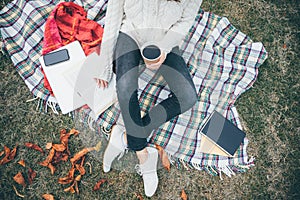 Student young hipster girl sitting on the blanket in autumn park garden reading book with a coffee to go cup in her hand