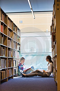Students reading book while sitting against bookshelf at library