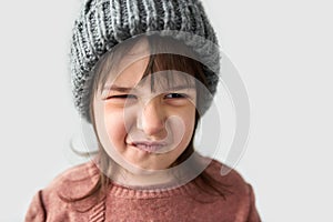 Studio closeup portrait of cute unhappy little girl with grumpy emotion in the winter warm gray hat, wearing sweater isolated on a