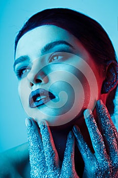 Studio fashion portrait of attractive woman with creative make-up and glitter and spangle on hands. Blue light.