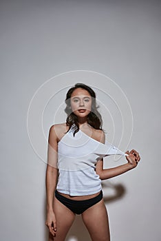 Studio fashion shot of provocative young woman in white shirt and panties looking at camera, adjusting shirt