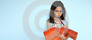 Studio horizontal image of a little girl wearing colorful dress, eyeglasses read a book posing on light blue background. Schoolkid