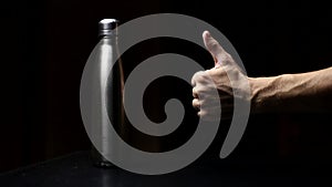 Studio image, close-up of male hand showing thumbs up about reusable, steel thermo water bottle on table, isolated on black backgr