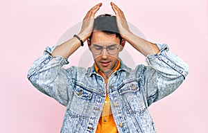 Studio image of a frustrated young man dressed in a denim jacket and transparent spectacles keeping both hands on his head