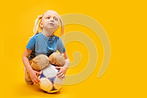 Studio photo of small caucasian white-haired girl with two tails holds soccer ball in isolation on yellow background. Copy space