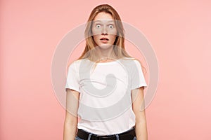 Studio photo of young amazed long haired redhead lady rounding surprisedly her green eyes while looking at camera,  over