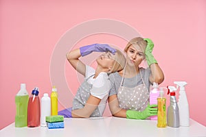 Studio photo of young blonde females dressed in basic t-shirts and aprons sitting over pink background with household chemicals,