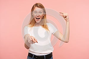Studio photo of young excited redhead female with natural makeup showing small size with raised hand and pointing emotionally at