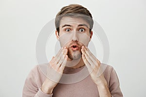 Studio portrait of amazed young man with bristle touching his face with surprised expression, standing against gray