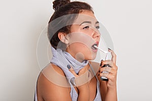 Studio portrait of attractive upset young brunette woman using spray to treat sore throat, female with scarf around neck. Throat